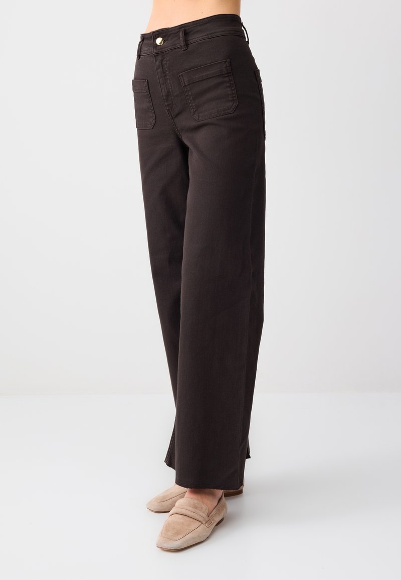 Personne portant un pantalon noir à jambes larges et des mocassins beige, debout devant un fond clair uni.