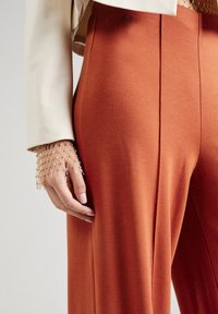 Rostfärgade byxor med vida ben i mjukt tyg, med rena linjer och hög midja, prydda med en hand dekorerad med ett armband av guldpärlor.