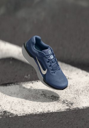 Chaussure de running Nike bleue avec logo swoosh blanc flottant au-dessus d’un bitume noir avec des marquages blancs peints.
