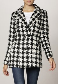 Doppelreihiger Mantel mit schwarz-weißem Hahnentrittmuster, mit Reverskragen und strukturierter Stoff. Verfügt über zwei schwarze Knöpfe an der Vorderseite.