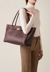 Braune Leder-Tote mit doppelten Griffen, mit einem goldfarbenen Verschluss und einer subtil strukturierten Oberfläche, die von einer Person in neutraler Kleidung gehalten wird.