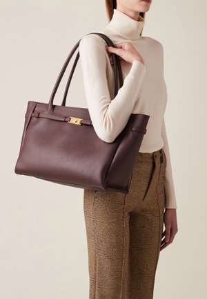 Brown leather tote bag with dual handles, featuring a gold-tone clasp and subtle textured surface, held by a person in a neutral outfit.