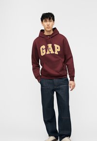 Νέος άνδρας που φοράει μπορντό φούτερ GAP, σκούρο τζιν και λευκά αθλητικά παπούτσια, στέκεται μπροστά σε ένα απλό φωτεινό φόντο.