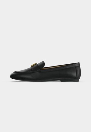 Chaussure mocassin en cuir noir avec un détail boucle en métal doré sur le dessus, semelle plate et couture le long du bord supérieur.