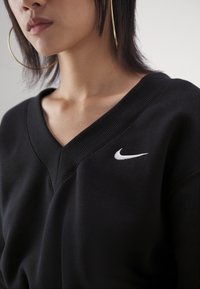 Svart sweatshirt med V-ringning, ribbad halsringning och en liten vit Nike-logotyp på vänster bröst. Mjuk tygtextur.