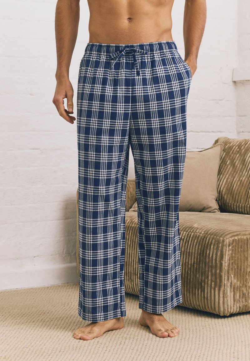 Shirtless man wearing blue and white plaid pajama pants, standing barefoot on carpeted floor next to beige corduroy sofa with cushions.