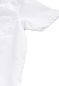 Gros plan d'un vêtement blanc à manches courtes montrant la texture du tissu et les détails de la couture de l'épaule sur un fond uni.