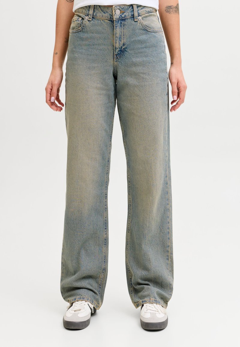 Person wearing loose-fitting faded blue jeans and white sneakers, standing with arms relaxed by their sides.