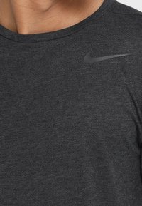 Camiseta deportiva de color gris oscuro con cuello redondo, mangas cortas y un pequeño logo reflectante de Nike en el hombro izquierdo. Textura suave.