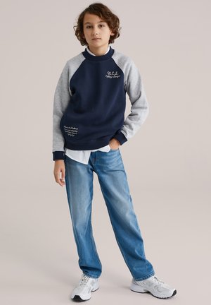 Felpa blu navy e grigia con maniche raglan, caratterizzata da testo ricamato. Abbinata a jeans azzurri e sneaker bianche.