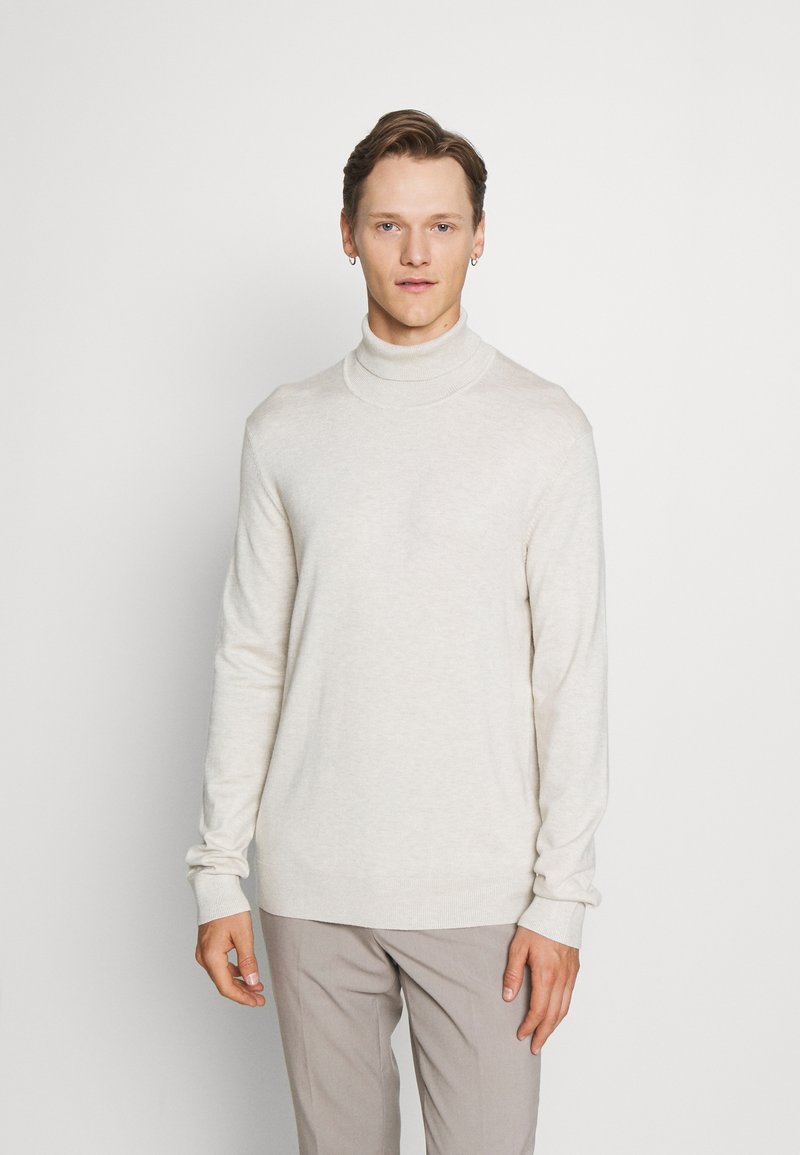Maglione a collo alto beige chiaro in morbido tessuto a maglia, con maniche lunghe e vestibilità rilassata, abbinato a pantaloni grigi chiari.