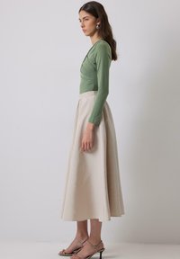 Forest green long-sleeve top with a ruched design paired with a flowing beige A-line skirt and light-colored strappy heeled sandals.