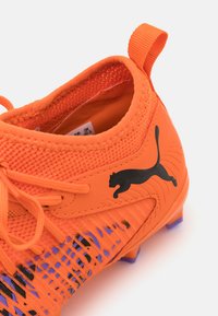 Fényes narancssárga focicipő, amely hálós felsőrésszel, fekete Puma logóval és texturált mintázatokkal rendelkezik a külső részen. Könnyű felhúzást biztosító húzófüllel van ellátva.
