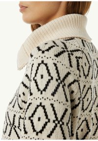 Sweter dzianinowy w kolorze kremowym z czarnymi wzorami geometrycznymi. Żebrowany kołnierz w jaśniejszym odcieniu kremu. Miękki, fakturowany materiał z wełny.