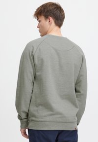 Sudadera gris con cuello redondo, mangas raglán, puños y dobladillo acanalados. Textura suave; detalle de logo en el lado inferior.