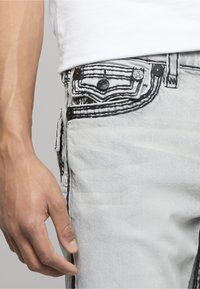 Jeans de mezclilla gris claro con costuras negras, con un diseño detallado en el bolsillo y detalles de botones, que muestran una textura suave y descolorida.