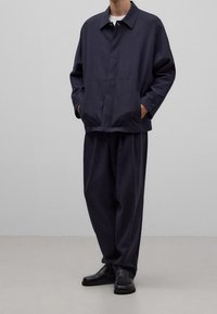 Chaqueta ligera azul marino con cierre de botones y dos bolsillos delanteros, combinada con pantalones sueltos azul marino y zapatos negros.