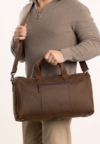 Bruine leren duffeltas met dubbele handvatten en een afneembare schouderriem. Heeft een voorvak met rits en met klinknagels versterkte accenten aan de onderzijde.