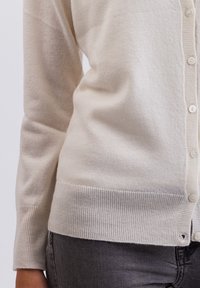 Ljusbeige kofta i mjuk ull. Har ribbade ärmar och nederkant, samt små knappar längst fram, med en slät yta.