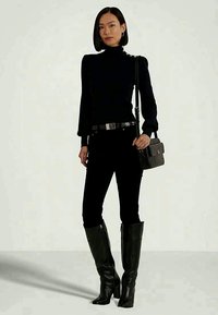 Ženska v črni obleki, sestavljeni iz oprijetega puloverja z visoko ovratnico, hlač z visokim pasom, škornjev s peto do kolen in torbice.