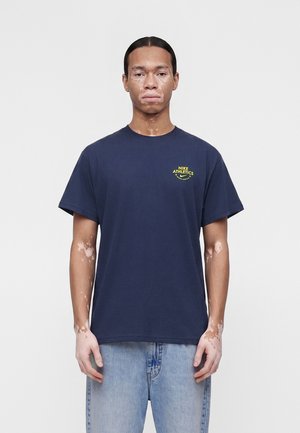 Moški z vitiligom, ki nosi temno modro majico Nike Athletics in svetlo modre kavbojke, stoji pred enobarvnim svetlo sivim ozadjem.