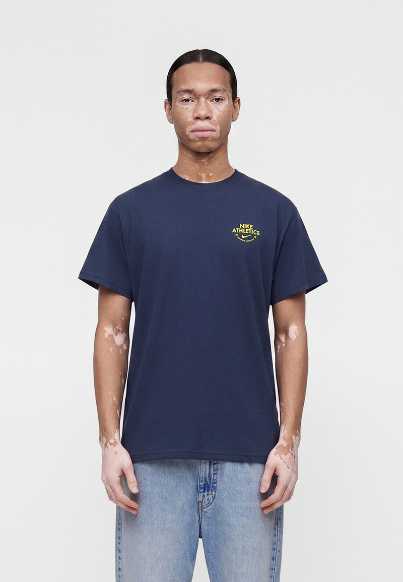 Uomo con vitiligine che indossa una t-shirt Nike Athletics blu navy e jeans azzurri chiari, in piedi davanti a uno sfondo semplice grigio chiaro.