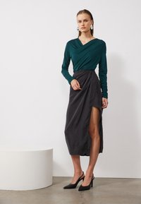 Teal long-sleeve top with a wrap design, paired with a dark gray skirt featuring a high slit. Model wears black pointed-toe heels.