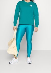 Turkusowa bluza, błyszczące turkusowe legginsy i białe buty. Trzyma jasnobeżową torbę. Posiada logo zarówno na bluzie, jak i na legginsach.