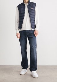 Navy puffer vest with horizontal stitching over a cream sweater, paired with blue jeans and white sneakers on a neutral background.