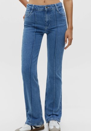 Jean bleu taille haute, coupe droite avec des détails de coutures verticales, porté avec des baskets blanches sur une personne debout sur un fond uni.