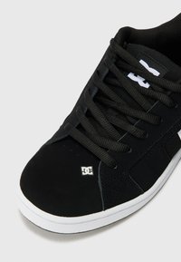 Sort sneaker med en glat ruskinds- og stofoverdel, med snøring foran, polstret krave og en hvid gummisål. DC-logoet prydes fremtrædende.