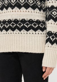 Strickpullover mit cremefarbener Basis und schwarzen geometrischen Mustern und Texturen. Gerippte Bündchen und Saum sorgen für Struktur; lange Ärmel sind sichtbar.