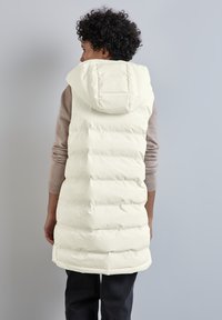 Krémový prošívaný vestě s polstrovaným designem, velkou kapucí a střední délkou. Vlastnosti zahrnují hladkou texturu a boční rozparky na spodní části.