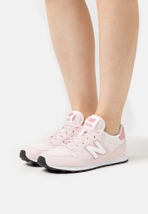 Piedi che indossano scarpe da ginnastica New Balance 500 rosa chiaro con logo bianco e suole nere, in piedi su una superficie bianca.