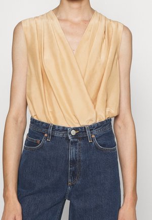 Ærmeløs beige satinviklebluse stoppet ned i højtaljede blå denimjeans på en person med neutral hudtone.