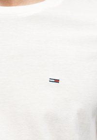 Λευκή κοντομάνικη μπλούζα με στρογγυλή λαιμόκοψη, με μικρό οριζόντιο ορθογώνιο λογότυπο με κόκκινες, λευκές και ναυτικές μπλε ρίγες στο στήθος.