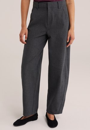 Femme portant un pantalon large gris foncé et des chaussures plates noires, debout les mains détendues le long du corps.