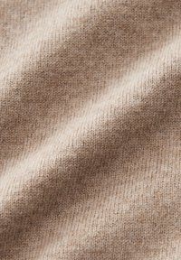 Nahaufnahme eines weichen, beigen Strickstoffs mit sichtbarer Textur und sanften Falten, die Schatten und Highlights erzeugen.