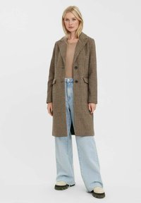 Hnědý kostkovaný dlouhý kabát s klopami, předními kapsami a dvěma černými knoflíky, nošený přes béžový top a světle modré široké džíny.