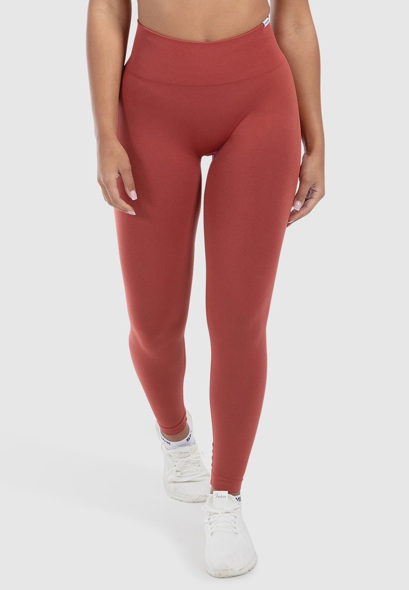 Leggings taille haute de couleur rouille en tissu extensible, présentant une texture lisse et une coupe fuselée, associés à des baskets blanches.