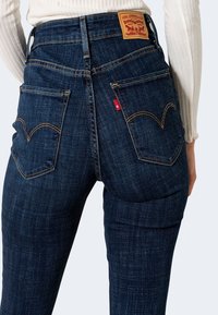Jeans de mezclilla oscuros de talle alto con un acabado texturizado, que presentan costuras amarillas distintivas y dos bolsillos traseros con un diseño curvado.