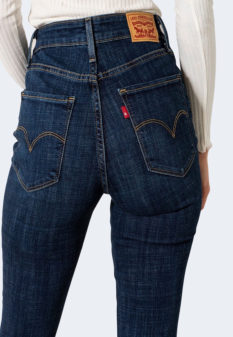 Jeans in denim scuro a vita alta con una finitura texturizzata, caratterizzati da cuciture gialle distintive e due tasche posteriori con un design curvo.