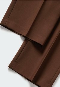 Jambes de pantalon marron sur mesure avec des plis nets, étendues à plat sur une surface blanche, montrant la texture du tissu et les détails de l'ourlet.