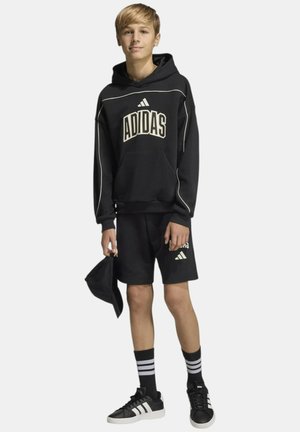 Ragazzo adolescente che indossa una felpa con cappuccio Adidas nera, pantaloncini Adidas neri, cappellino Adidas nero in mano, calzini neri con strisce bianche e scarpe da ginnastica nere.