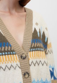 Gros plan sur une personne portant un cardigan beige avec des motifs géométriques bleus, marron et jaunes, et deux grands boutons à l'avant.