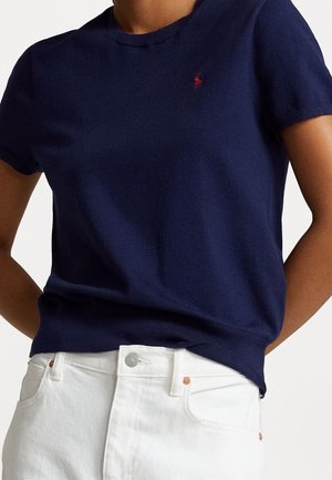 Personne portant une chemise bleu marine à manches courtes avec un petit logo rouge sur la poitrine, associée à un pantalon blanc taille haute.