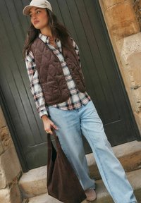 Junge Frau mit beigem Cap, brauner gesteppter Weste über einem karierten Hemd, hellblauen Jeans, die eine große braune Wildleder-Handtasche hält und an einer Steintreppe steht.