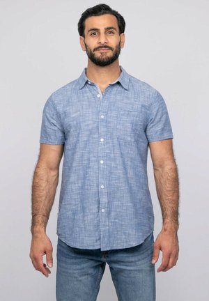Homme aux cheveux foncés et à la barbe, portant une chemise bleu clair à manches courtes boutonnée et un jean, debout devant un fond gris uni.