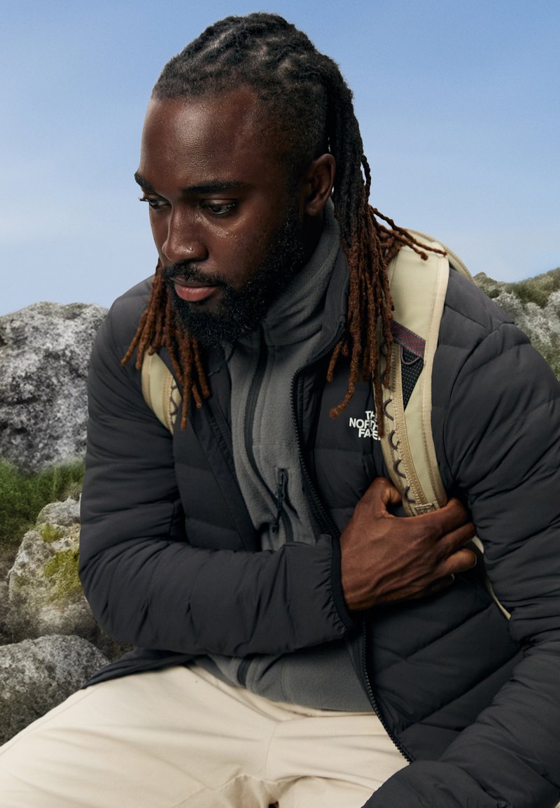 Casaco puffer preto com textura acolchoada, sobre um fleece cinza com fecho. Alça de mochila bege visível. Modelo sentado no chão rochoso.