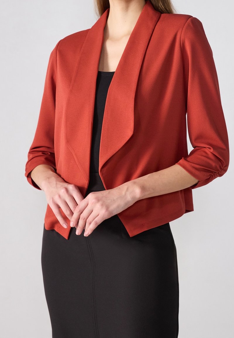 Blazer rojo cortado con cuello chal, mangas tres cuartos y textura suave, combinado con un vestido negro ajustado.